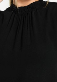 Blouse noire à col montant avec un détail de tissu froncé autour du col et des mèches de cheveux blonds clairs visibles sur le dessus.