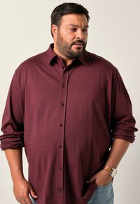 Uomo con barba che indossa una camicia a maniche lunghe color maroon arrotolata e un orologio, guarda in basso e di lato con una mano in tasca.