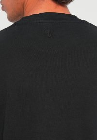 De nek en schouder van een man die een zwart gebreid bovenstuk draagt met een klein geborduurd logo centraal onder de halslijn op de rug.