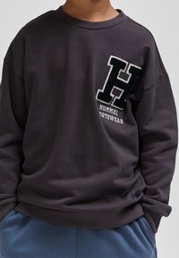 Ciemnoszara bluza z okrągłym dekoltem, luźnym krojem, z teksturowaną czarną naszywką w kształcie litery "H" oraz białym napisem "HUMMEL SPORTSWEAR" z przodu.
