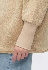 Μπεζ φλις φούτερ με υφή στην επιφάνεια και μπεζ μανσέτες με ριμπ σχέδιο, σε συνδυασμό με ανοιχτό μπλε τζιν τζιν. Κοντινή λήψη στο μανίκι και το χέρι.