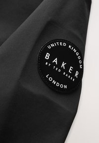 Čierna látka s okrúhlym čiernym štítkom označeným "United Kingdom Baker by Ted Baker London" prišitým na ňu.