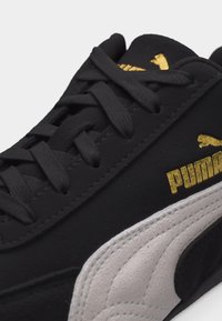 Μαύρο αθλητικό παπούτσι Puma με γκρι σουέντ ρίγα και χρυσό λογότυπο Puma στο πλάι, με μαύρα κορδόνια και λεπτομερείς ραφές.