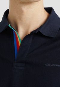 Närbild på en person som bär en marinblå pikétröja med en flerfärgad randig placket och Karl Lagerfeld-logotyp på bröstet.