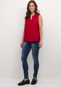 Piros ujjatlan felső kerek nyakkivágással és redőzéssel, sötétkék skinny farmernadrággal és fekete loaferekkel párosítva. Egyszerű háttér.