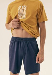 Żółta bawełniana koszulka z grafiką "Sunrise Surf Club" w białym kolorze, zestawiona z ciemnogranatowymi szortami z elastycznym pasem i boczną kieszenią.