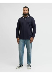 Marineblaues Langarm-Poloshirt mit hellfarbigem Kragen, kombiniert mit hellblauen Jeans und schwarzen Sneakers, vor einem neutralen Hintergrund.