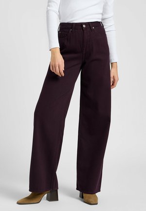 Personne porte un pantalon taille haute à jambes larges de couleur violet foncé avec des bottines à talons couleur beige et un haut blanc à manches longues, debout devant un fond uni.