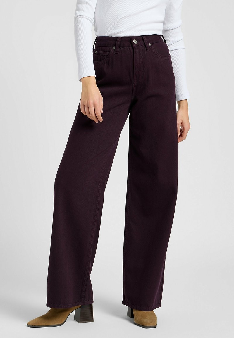 Personne porte un pantalon taille haute à jambes larges de couleur violet foncé avec des bottines à talons couleur beige et un haut blanc à manches longues, debout devant un fond uni.