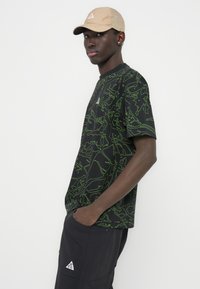 Μαύρο κοντομάνικο μπλουζάκι με πράσινο αφηρημένο μοτίβο γραμμών· διαθέτει μικρό λευκό λογότυπο στο στήθος. Συνδυάζεται με σκούρο, φαρδύ παντελόνι.