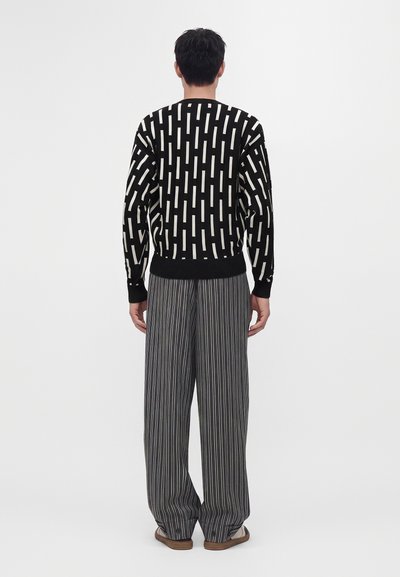 Homme debout, dos à l'appareil photo, portant un pull noir avec des rectangles blancs verticaux et un pantalon large gris à fines rayures.