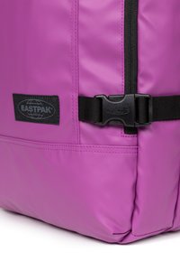 Fioletowy plecak Eastpak z gładką, matową fakturą, czarnymi elementami metalowymi, wyraźną naszywką z logo oraz zapięciem na klamrę z boku.