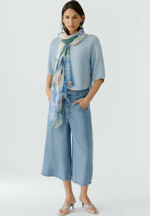 Frau trägt hellblaues kurzärmeliges Strickoberteil, weite Jeanshose, floralen Schal und silberne Sandalen mit Absatz, steht mit den Händen in den Taschen.