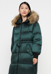 Abrigo tipo puffer verde con capucha con ribete de piel, cierre frontal con cremallera, cintura ajustable y bolsillos laterales. Presenta una textura brillante y acolchada.