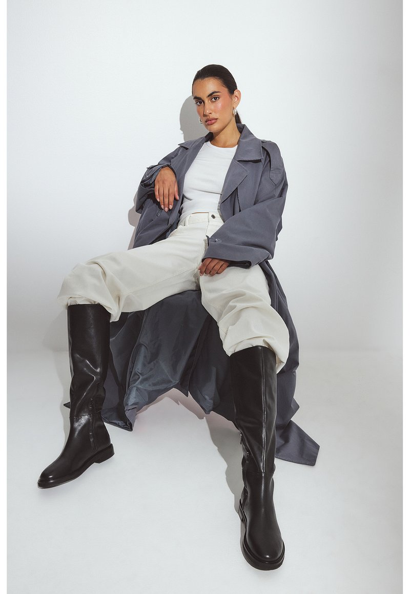 Grijze trenchcoat met een oversized pasvorm, witte t-shirt, wijde lichte broek en kniehoge zwarte laarzen; soepel materiaal en strakke lijnen.