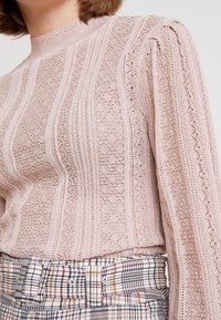 Jasnoróżowa dzianinowa bluzka z pionowymi wzorami, wysoki dekolt i bufiaste rękawy, w połączeniu z spodniami w kratę o wysokim stanie.