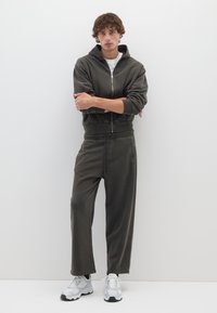 Felpa con cappuccio di colore verde oliva scuro con zip anteriore e tasca a marsupio, abbinata a pantaloni della tuta con coulisse dello stesso colore. Modello indossa una maglietta bianca e sneakers.