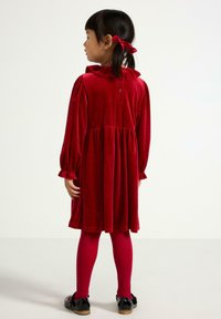 Czerwona sukienka z weluru z długimi rękawami, falbaniastym kołnierzykiem i marszczoną talią; zestawiona z różowymi rajstopami i czarnymi butami. Widok z tyłu.