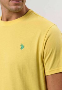 Kollane puuvillane t-särk, millel on ümmargune kaelus ja lühikesed varrukad. Rinnal on väike türkiissinine tikitud logo. Sile tekstuur.