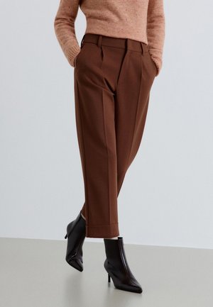 Persona che indossa pantaloni marroni plissettati, un maglione beige e stivaletti neri a punta, in piedi con le mani in tasca.
