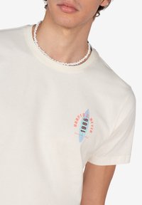 Camiseta de algodón crema con cuello redondo, mangas cortas y un gráfico de tabla de surf en rojo y azul con el texto "Harper 1985".