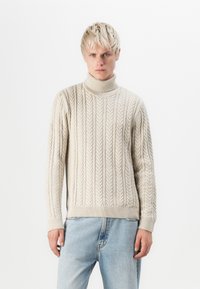Maglione a collo alto color crema con motivo a treccia, polsini e orlo a costine, abbinato a jeans in denim chiaro. La texture appare morbida e accogliente.
