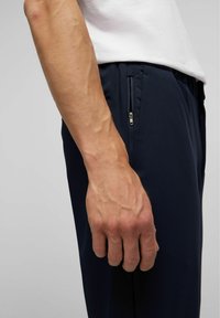 Marineblauwe sportbroek heeft een ritssluitingzak aan de zijkant. De stof lijkt glad, en de tailleband is elastisch voor een flexibele pasvorm.