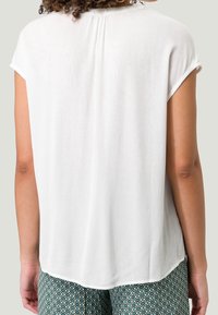 Top blanco de manga corta con un patrón texturizado, que presenta un dobladillo redondeado y un detalle de pliegue en la parte trasera. Combinado con pantalones verdes estampados.