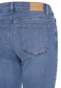 Helevaat denimpükste tagakülg helerohelises toonis, kus on üks tagatasku oranži õmblusega ja pruunist nahast õlipõhise kaubamärgi silt.