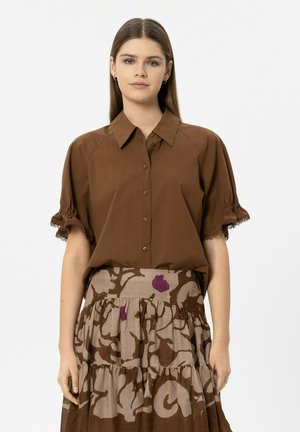 Young woman wearing a brown button-up shirt with ruffled sleeves and a beige skirt with brown and purple abstract floral patterns.