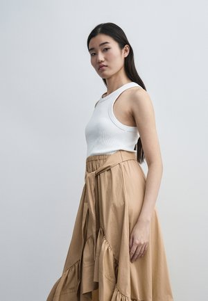 Mladá žena s dlhými tmavými vlasmi nosiaci biele tielko bez rukávov a vysoký pískový krátky rukáv so záhybmi, stojaca proti jednoliatemu pozadiu.