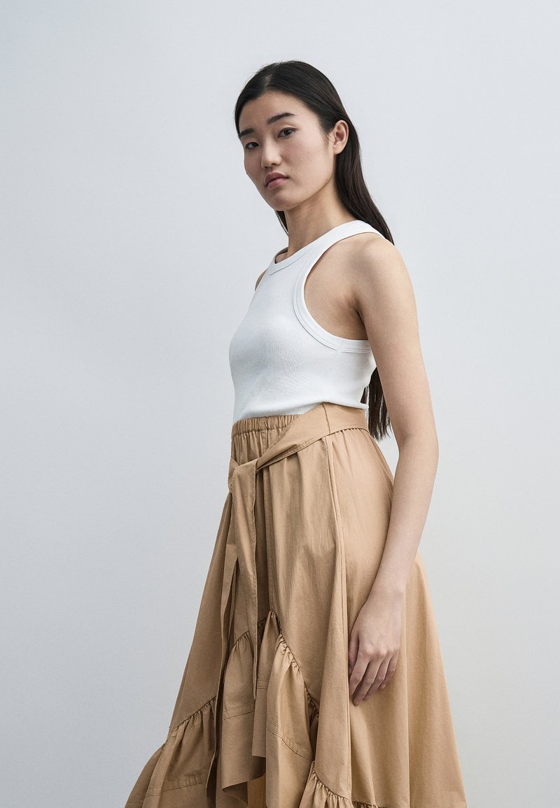 Νεαρή γυναίκα με μακριά σκούρα μαλλιά που φοράει λευκό αμάνικο τοπ και ψηλόμεση καφέ φούστα με βολάν, στέκεται μπροστά σε απλό φόντο.