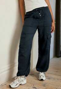 Pantaloni cargo navy con vita elastica e lacci, dotati di numerose tasche, polsini affusolati e una vestibilità rilassata. Abbinati a sneakers grigie.