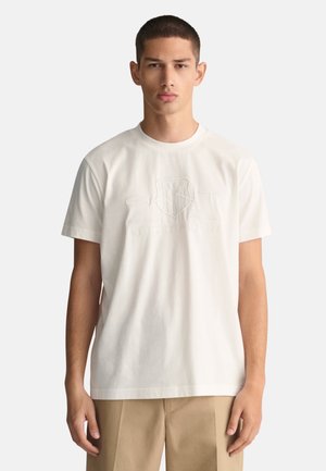 Light beige cotton t-shirt with a round neckline, featuring embossed brand logo and text on the front. Short sleeves and relaxed fit.
