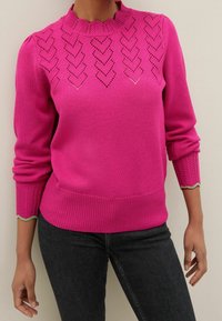 Woman wearing a bright pink knitted sweater with heart-shaped patterns across the chest and scalloped green trim on the cuffs, paired with black jeans.