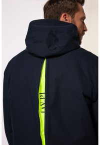 Laivastonsininen vedenpitävä takki hupulla, jossa on keskellä neon keltainen vetoketju ja painettu bränditys takana, valmistettu sileästä kankaasta.