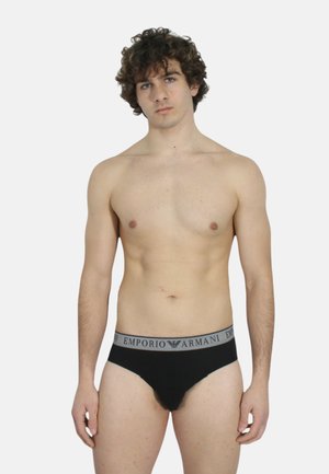 Slip in cotone nero con elastico in vita grigio Emporio Armani, caratterizzati da una vestibilità aderente e una texture liscia. Il modello è a torso nudo e sta in piedi.