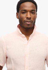 Jasnoróżowa lniana koszula z krótkim rękawem i kołnierzykiem mandaryńskim z guzikami. Posiada małe haftowane logo na piersi. Gładka tekstura.