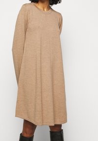 Robe en tricot beige à manches longues, décolleté rond et coupe ample. Présente une texture subtile et un ourlet droit. Portée avec des bottes noires montantes.