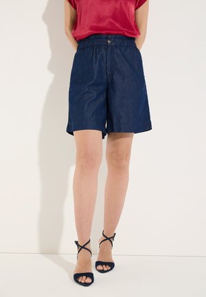 Persoon draagt donkerblauwe shorts met hoge taille, rood topje en marineblauwe sandalen met hak en bandjes, staand tegen een witte muur.