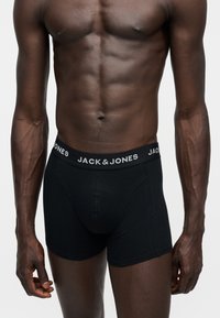 Mužský trup v čiernych boxerkách Jack & Jones s logom na páse, ukazujúci svalnaté a žilnaté ruky na jednofarebnom pozadí.