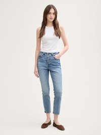 Kvinna som står med ena handen i fickan, iklädd vit ärmlös topp, ljusblå jeans och bruna platta skor mot beige bakgrund.