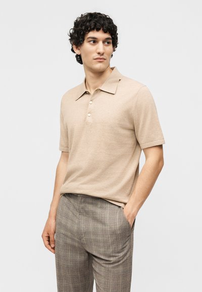 Bărbat cu păr creț, purtând un tricou polo bej cu mâneci scurte și pantaloni gri cu carouri, stând cu o mână în buzunar.