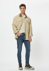 Joven vestido con una chaqueta oversized beige, una camisa beige clara, unos vaqueros azules ajustados y zapatillas verdes de caña alta, de pie frente a un fondo blanco.