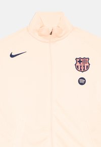 Nike full-zip jas in licht perzik met hoge kraag, marineblauw logo op de linker borst en het FC Barcelona-embleem. Huidige textuur, twee zijzakken.