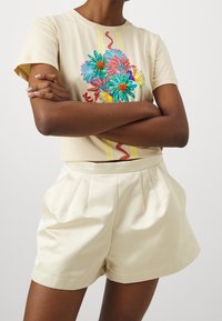 Camiseta de algodón beige con bordado floral multicolor y rayas verticales, combinada con pantalones cortos crema de cintura alta con pliegues.