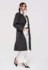 Femme portant un long trench-coat noir par-dessus une chemise blanche boutonnée et un pantalon blanc à jambes larges, avec des talons hauts noirs, posant de profil.