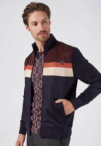 Navy zip-up jacket with maroon and cream stripes across the chest. Features side pockets and ribbed cuffs. Worn over a patterned shirt.
