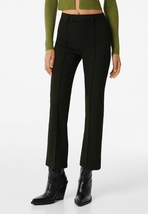 Femme portant un pantalon noir sur mesure, des bottines à talons noires et un pull vert court, montrée de la taille aux pieds.