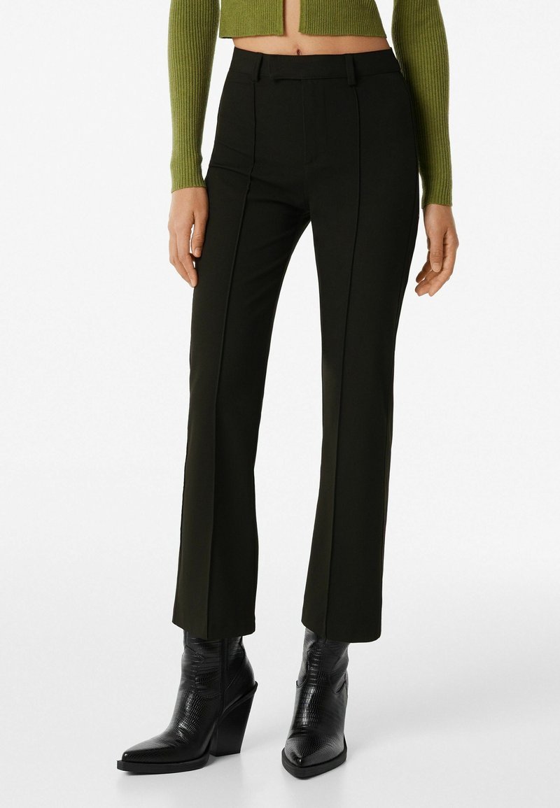 Femme portant un pantalon noir sur mesure, des bottines à talons noires et un pull vert court, montrée de la taille aux pieds.